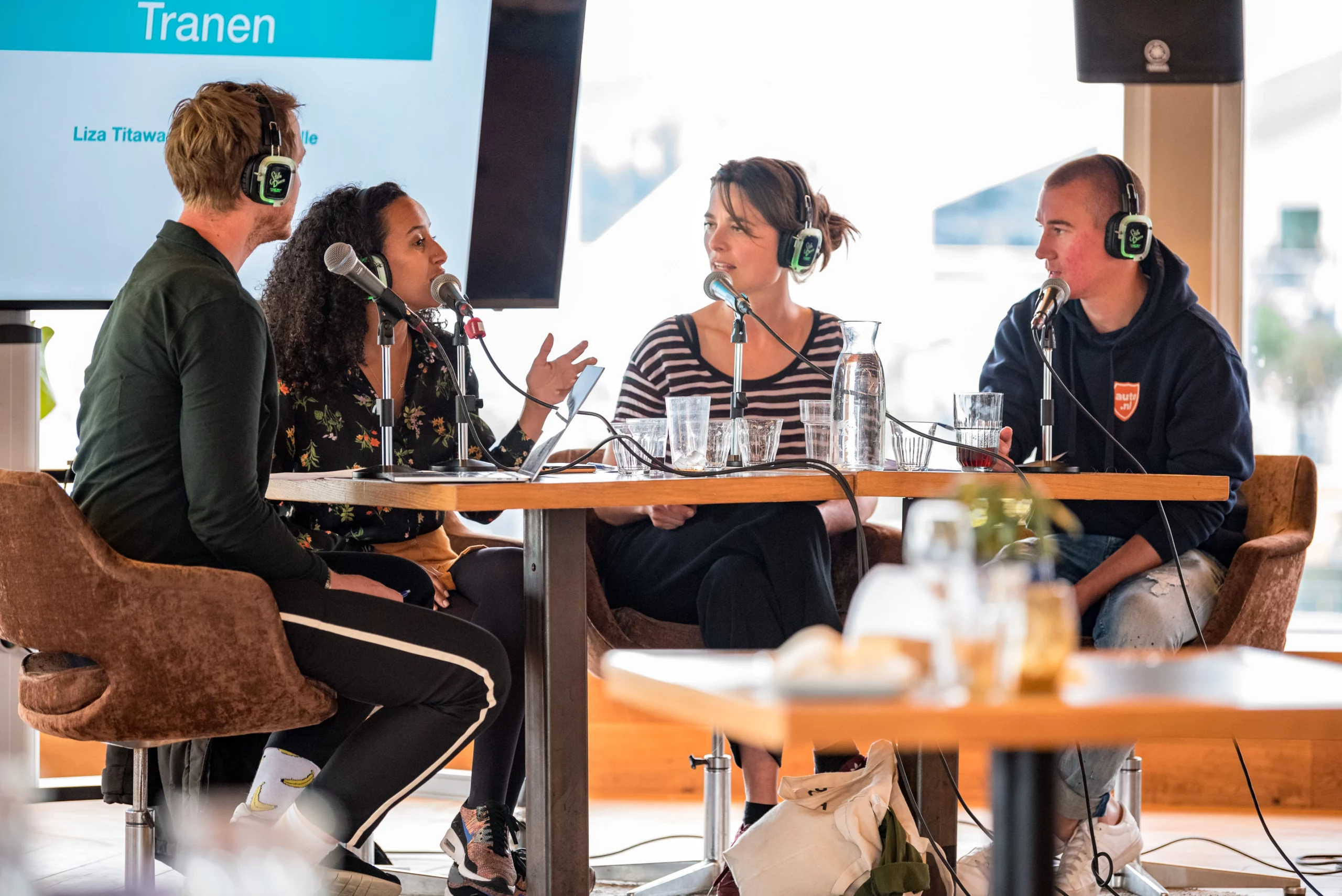 Vier personen zitten aan een tafel met microfoons en koptelefoons tijdens een live podcastopname. Op de achtergrond is een scherm zichtbaar met de tekst 'Tranen' en de naam 'Liza Titawano'