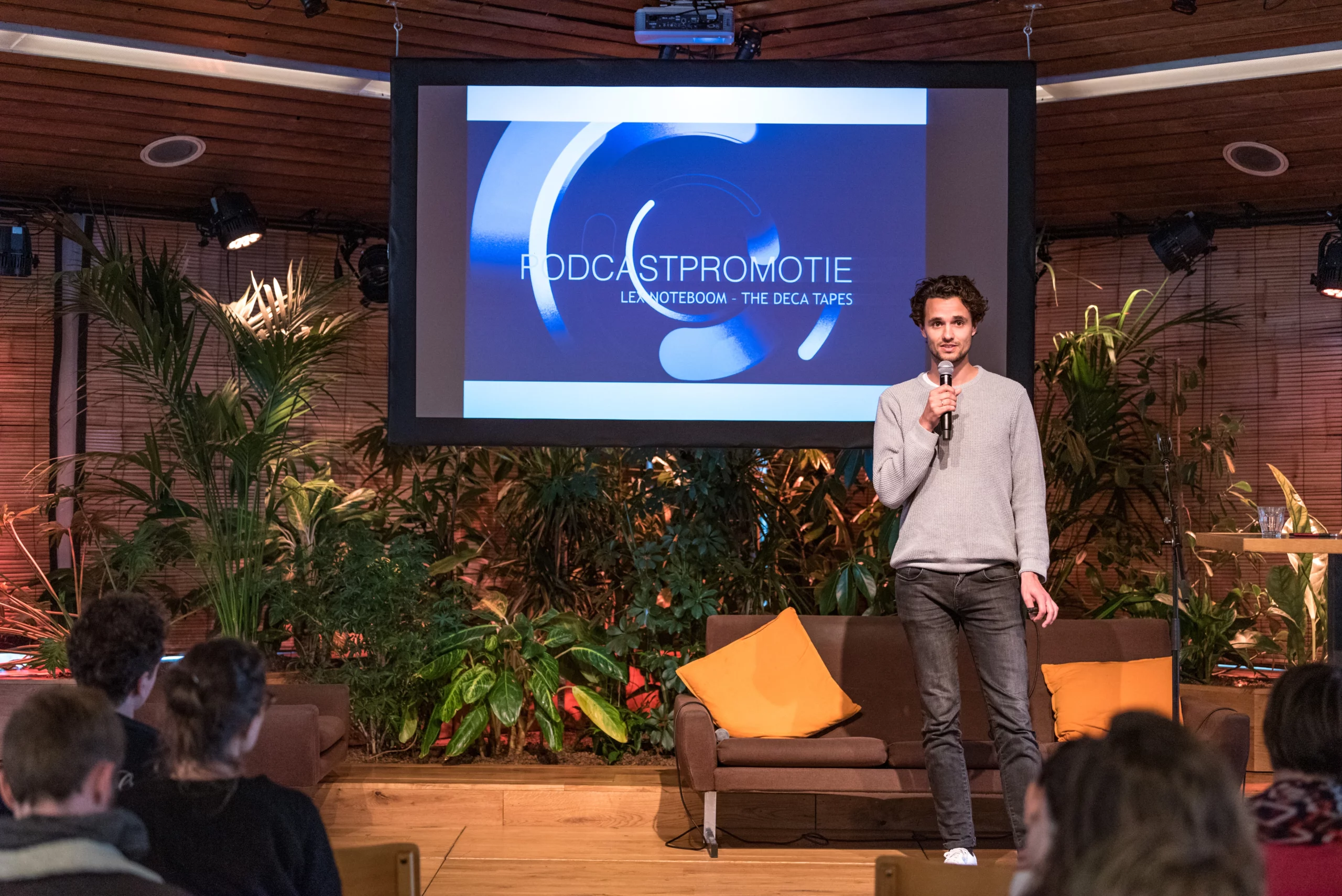 Een man staat op een podium met een microfoon en presenteert voor een publiek. Achter hem is een groot scherm met de tekst 'Podcastpromotie' en 'Lex Noteboom – The Deca Tapes', omringd door groene planten