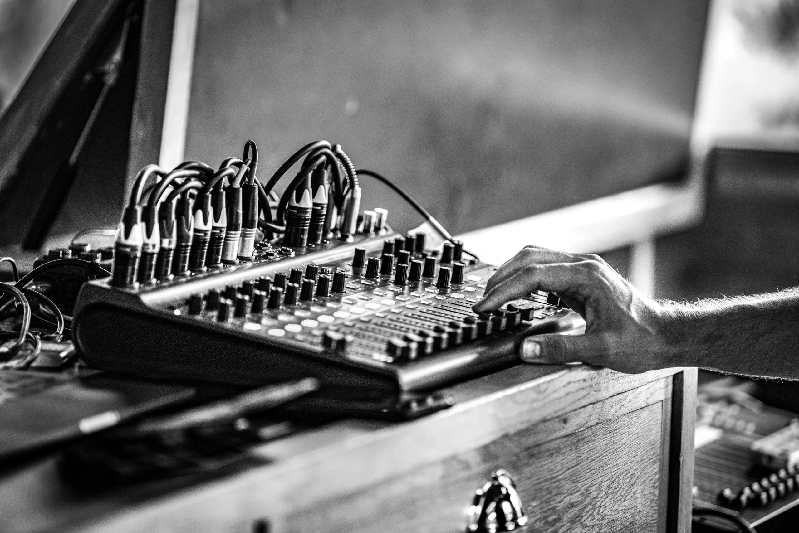 Een persoon bedient een geluidsmengpaneel met vele knoppen, schuifregelaars en aansluitkabels in zwart-wit.