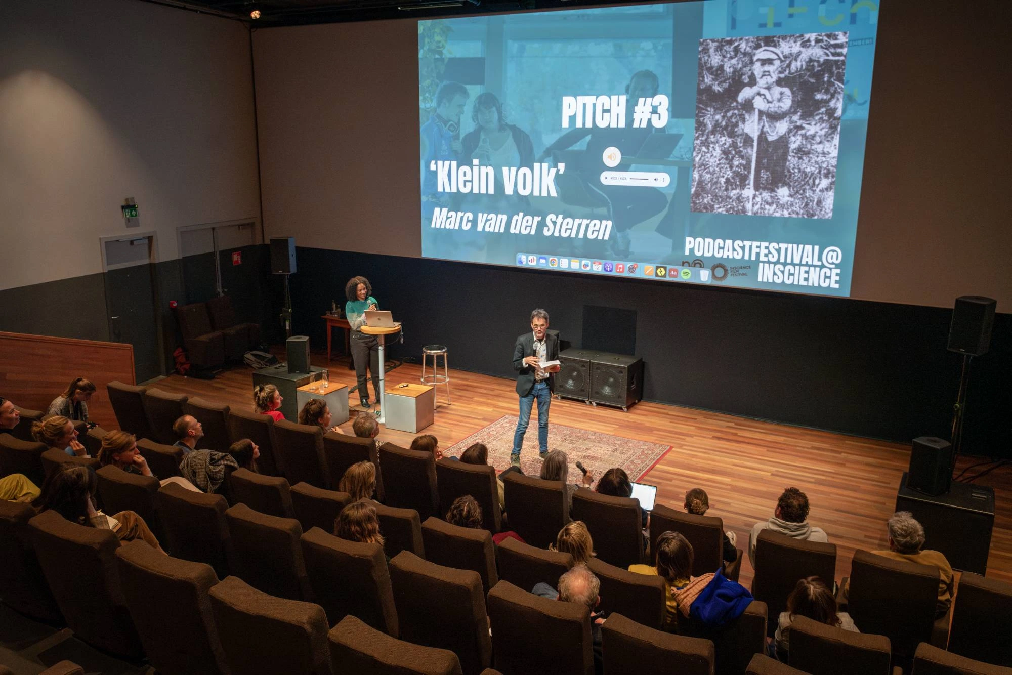 Twee mensen staan op een podium tijdens een live presentatie op het Podcastfestival@Inscience. Achter hen is een groot scherm zichtbaar met de tekst 'PITCH #3 Klein volk' en de naam 'Marc van der Sterren'. Het publiek zit in een zaal en luistert aandachtig
