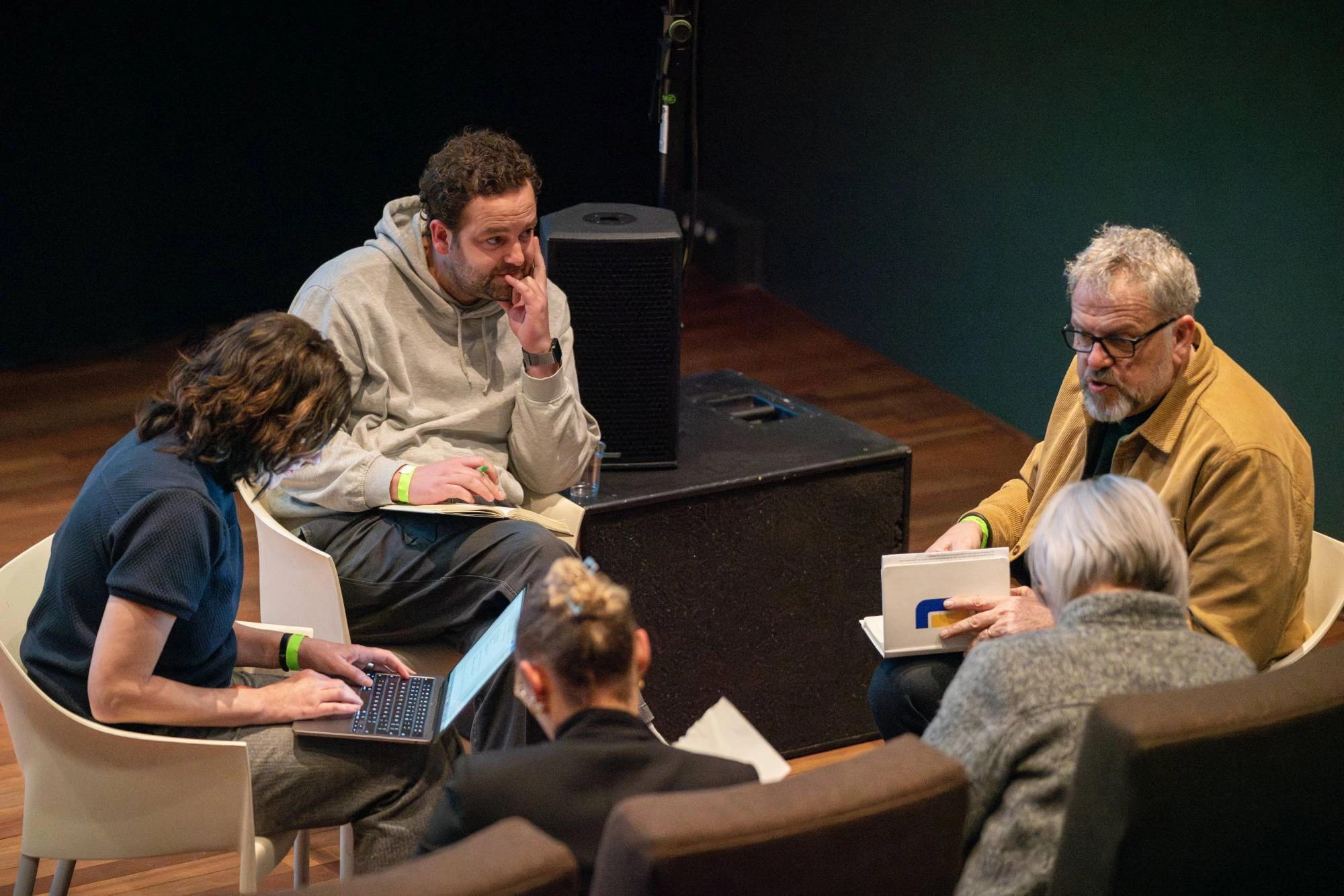 Vijf mensen zitten in een informele kring op het podium tijdens een live gesprek of paneldiscussie. Twee mannen zijn in gesprek, terwijl de anderen op laptops werken of luisteren