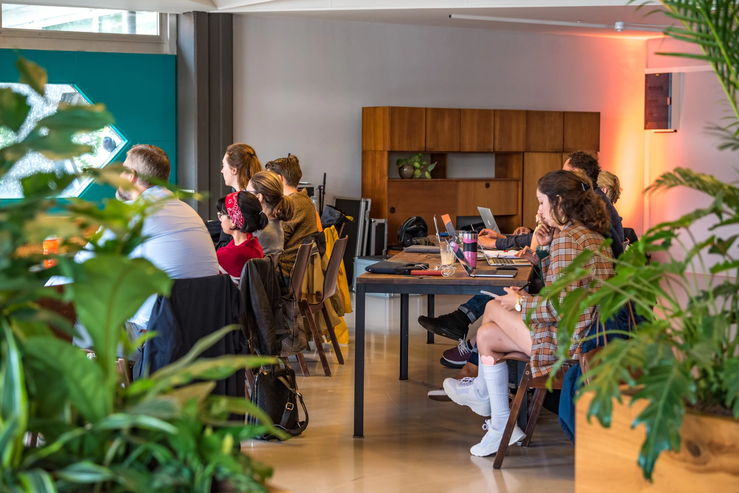 Groep mensen zit aan een lange tafel in een moderne, lichte ruimte met planten. Ze werken op laptops en volgen geconcentreerd een presentatie of vergadering.