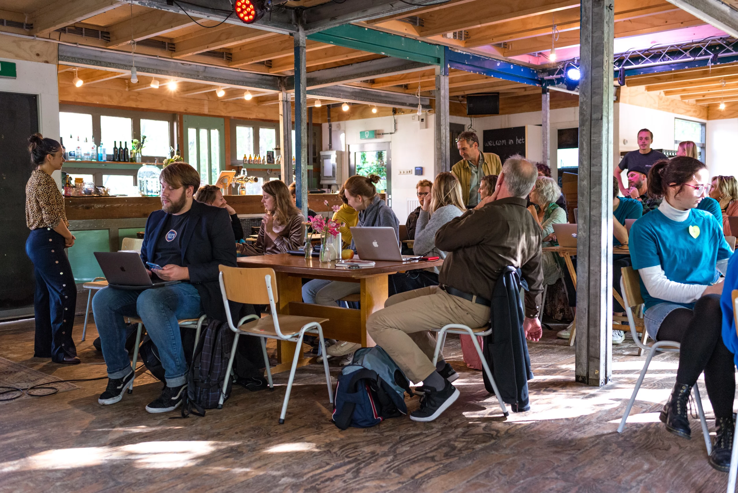 Groep mensen in een informele werk- of studieruimte met laptops, zittend aan tafels en in gesprek met elkaar. De ruimte heeft een ontspannen sfeer met houten vloeren, tafels en een bar op de achtergrond.