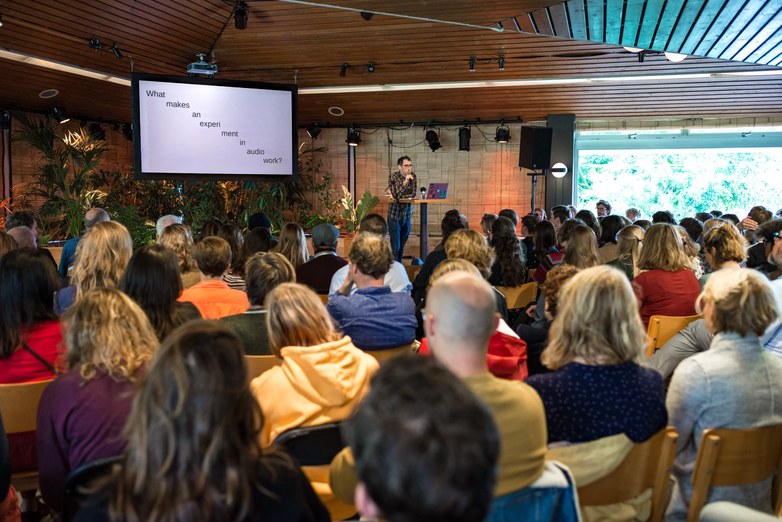 Een spreker staat voor een groot publiek in een zaal met houten wanden en veel planten. Op het scherm achter de spreker staat de tekst 'What makes an experiment in audio work?'. Het publiek zit op stoelen en luistert aandachtig.