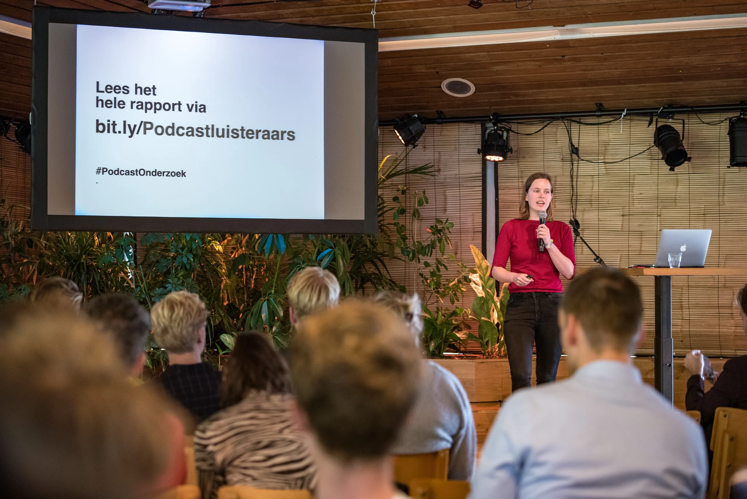 Een jonge witte vrouw met lang bruin haar staat op een podium met een microfoon en presenteert voor een publiek. Achter haar is een scherm zichtbaar met de tekst 'Lees het hele rapport via bit.ly/Podcastluisteraars #PodcastOnderzoek' en er staan planten langs de muur