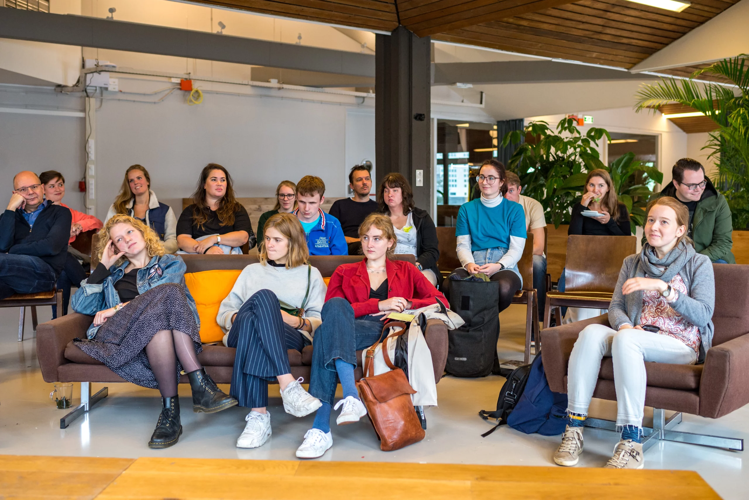 Een groep mensen zit in een informele kringen op stoelen en banken in een lichte, moderne ruimte met planten. Ze volgen aandachtig een presentatie of lezing