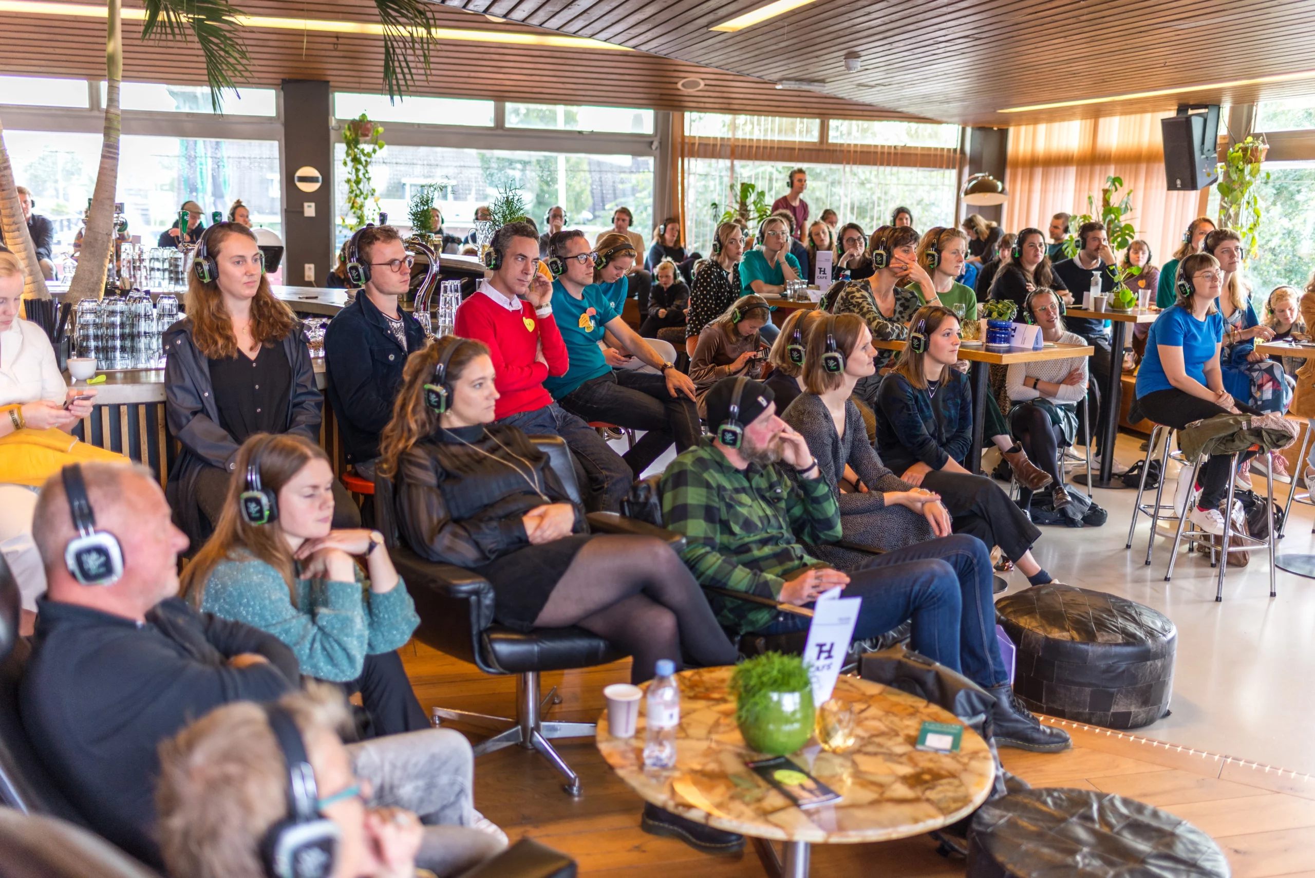 Een groep mensen zit in een moderne ruimte met grote ramen en planten. Ze dragen koptelefoons en luisteren geconcentreerd naar een audio-ervaring of presentatie