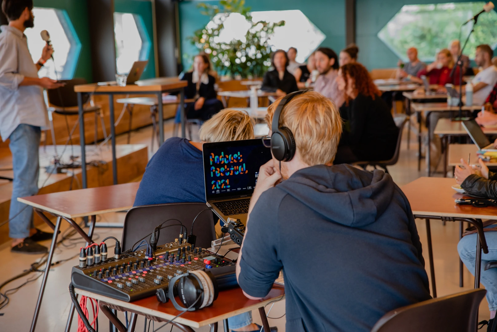 Twee personen met koptelefoons zitten achter een geluidsmengpaneel en een laptop tijdens een live opname of podcastregistratie. Op de achtergrond is een groep mensen in een zaal te zien die luistert en aantekeningen maakt