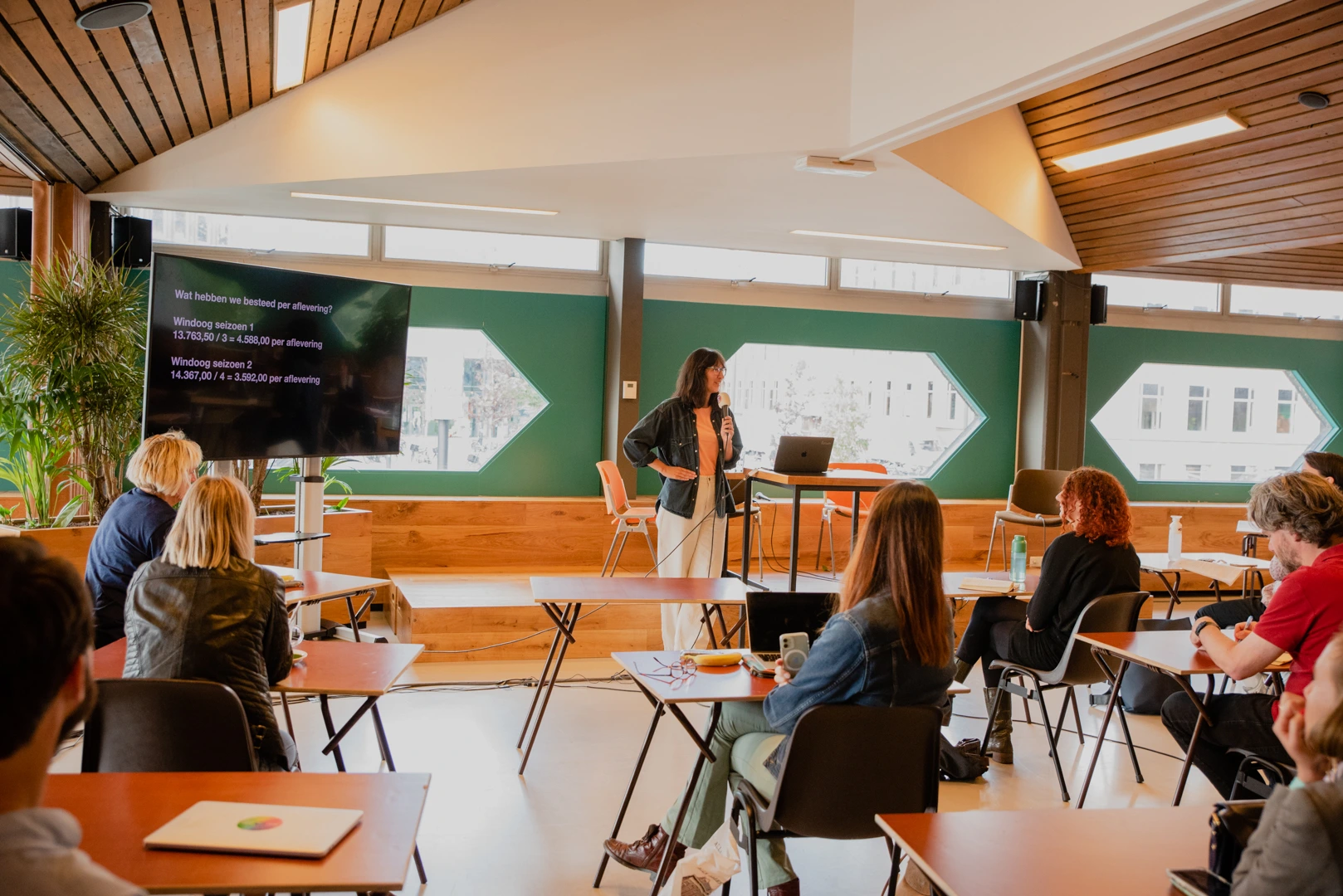 Een vrouw staat voor een groep mensen en presenteert met een microfoon in een lichte, moderne ruimte. Achter haar is een scherm met statistieken over Windoog en het aantal bestede uren per aflevering. Het publiek zit aan tafels en luistert aandachtig.