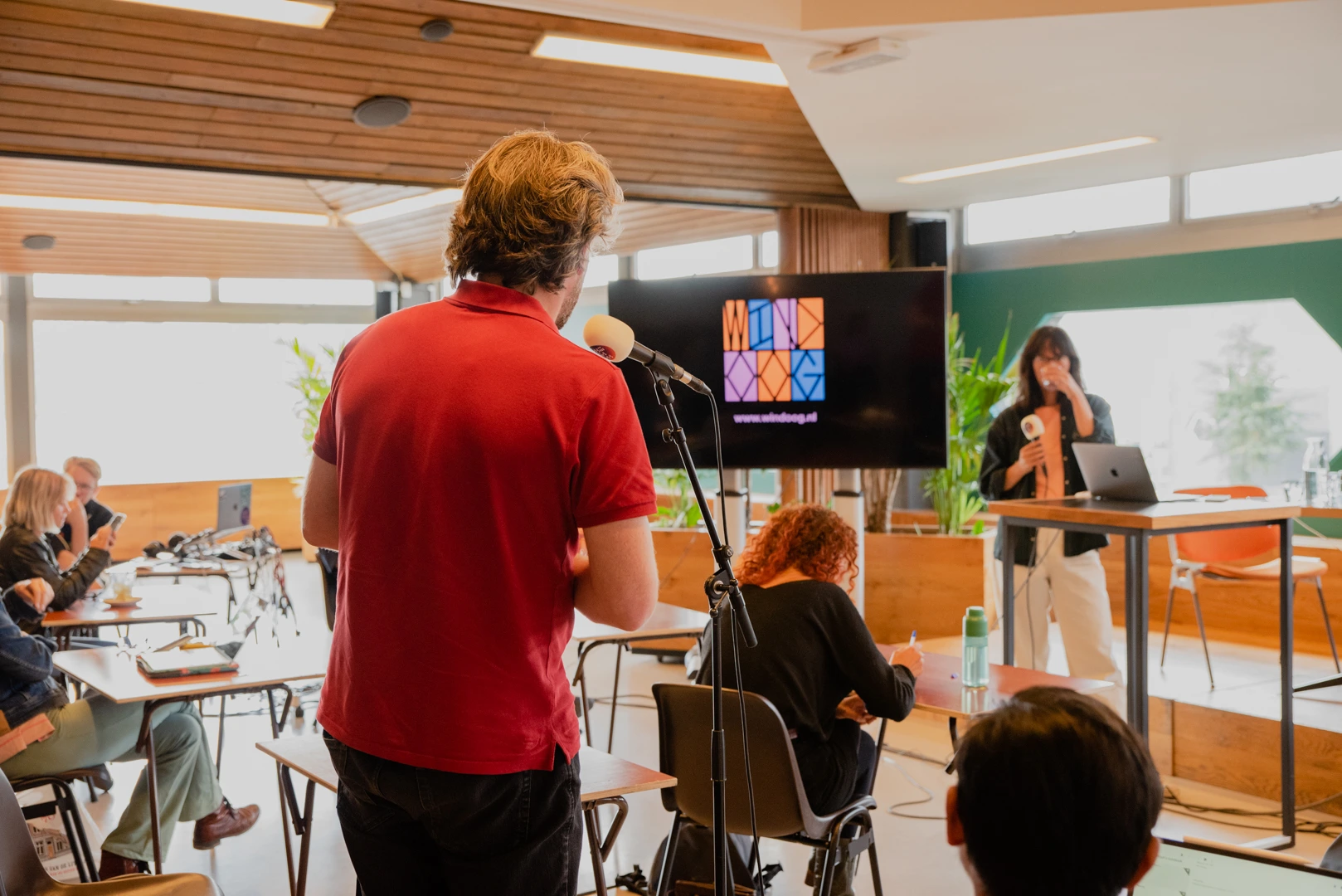 Een man in een rood shirt staat met zijn rug naar de camera en spreekt in een microfoon tijdens een live podcastopname. Achter hem zit een vrouw aan een tafel met een laptop en een koptelefoon. Op de achtergrond is een scherm met een logo en een websiteadres zichtbaar.