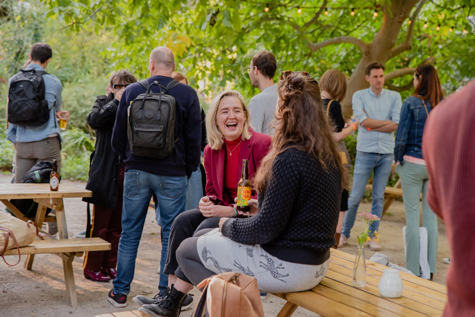Een groep mensen geniet van een informele buitenborrel in een groene omgeving. Twee vrouwen zitten lachend aan een houten picknicktafel met een flesje drank, terwijl anderen staan en met elkaar in gesprek zijn