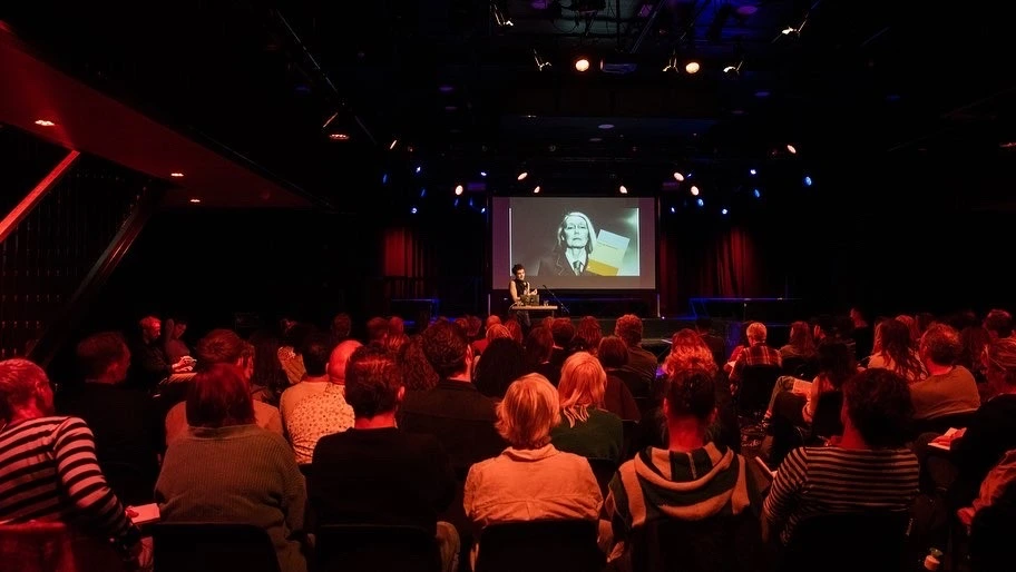 Een spreker staat op een podium in een donkere zaal met een groot scherm achter zich. Het publiek zit in rijen stoelen en luistert aandachtig naar de presentatie