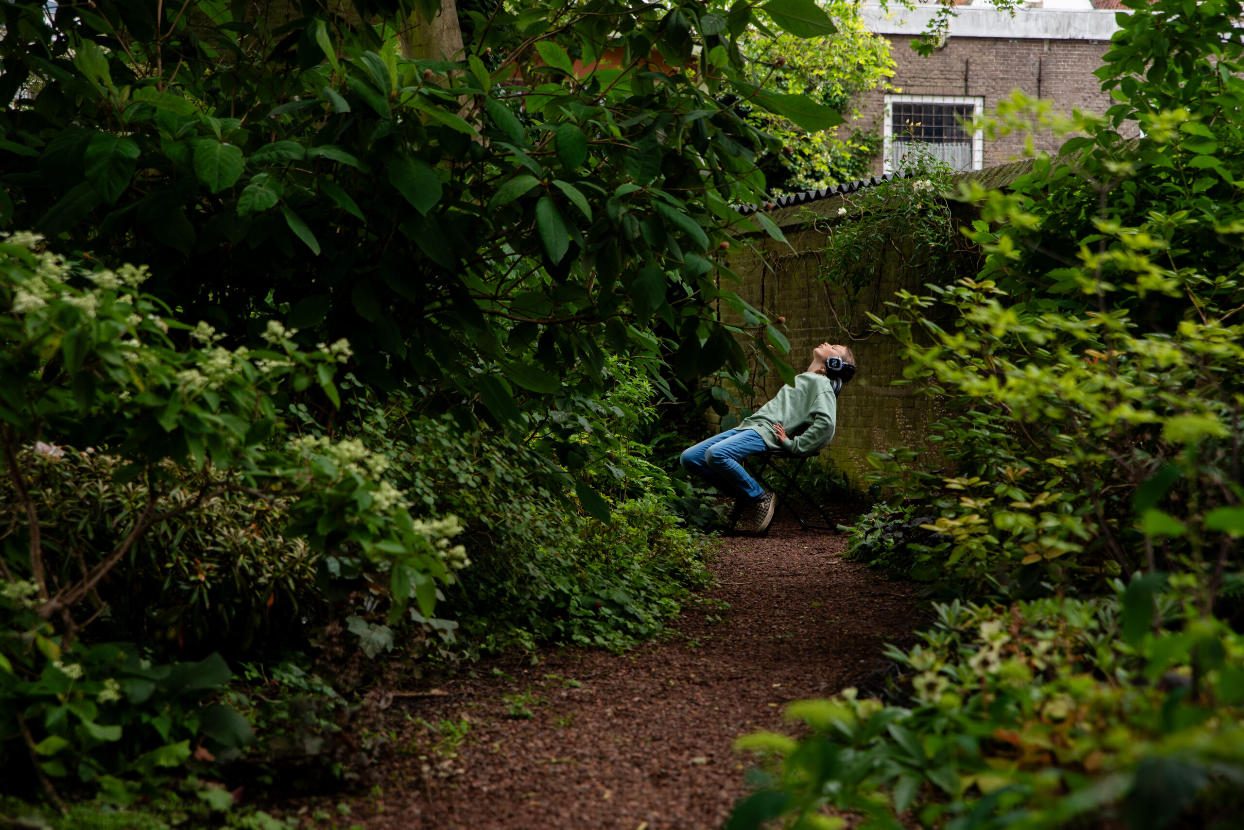 Een persoon zit in het midden van een weelderige groene tuin op een klein klapstoeltje. De persoon draagt een lichtgroene trui, een spijkerbroek en een grote zwarte over-ear koptelefoon. De persoonl leunt ver naar achteren met hun gezicht omhoog gericht naar de bomen en de lucht.