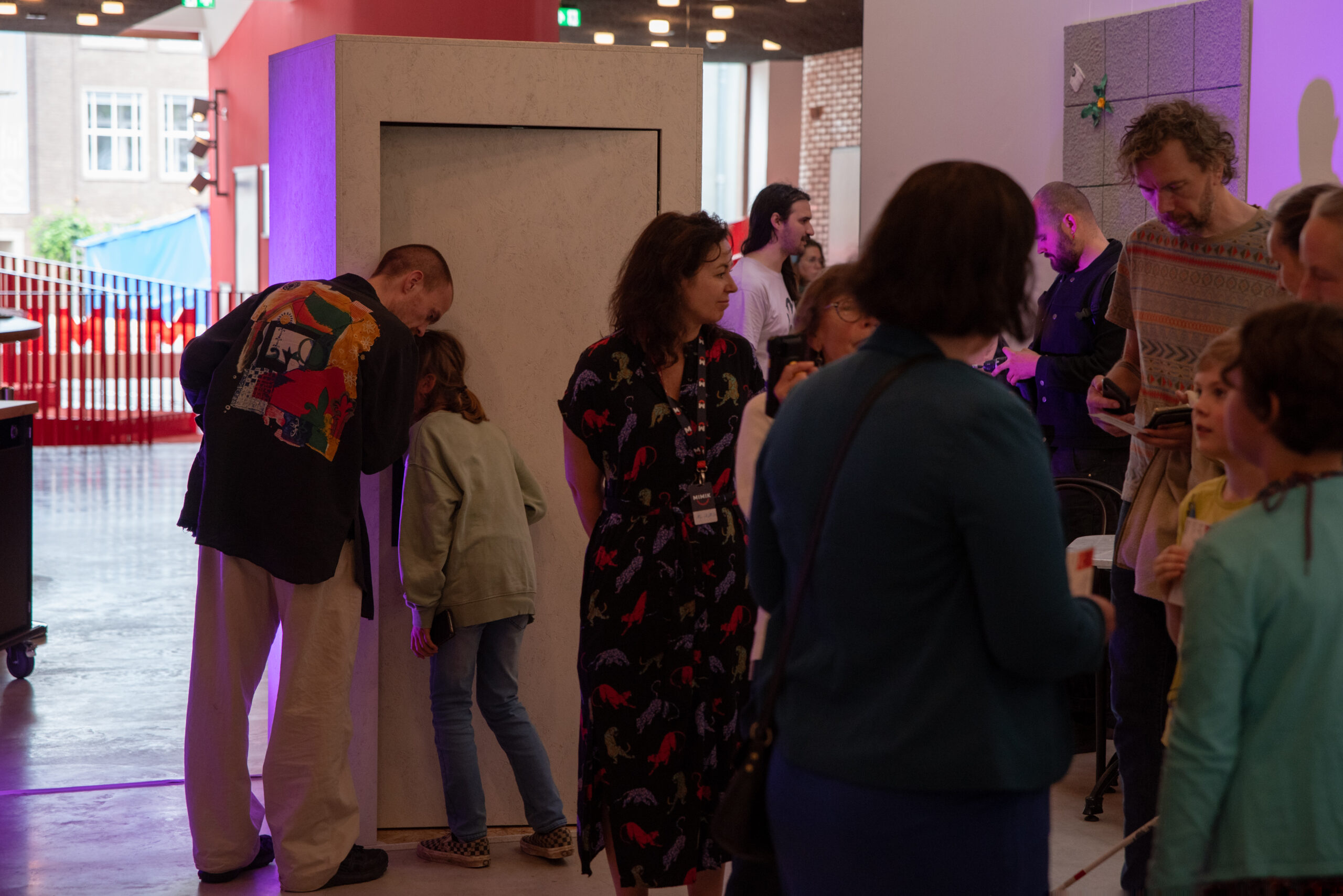 Een groep mensen in een foyer van een theater. Aan de linkerkant staan een man met een kleurrijk jasje en een kind in een groene trui voorovergebogen om in een hoge, witte cabine te kijken die verlicht is met paars licht. Aan de rechterkant staan verschillende andere bezoekers, waaronder een vrouw in een donkere jurk met een kleurrijke dierenprint, te praten of op hun telefoon te kijken.