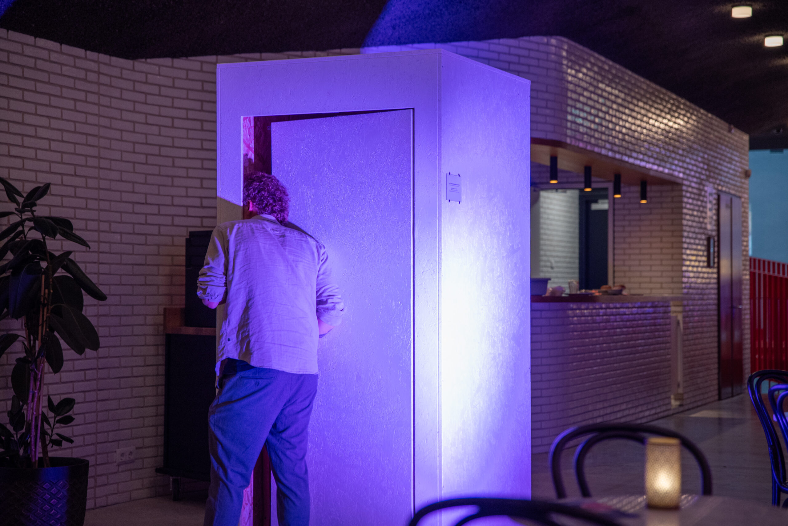 Een persoon op de rug gezien, met krullend haar, een licht overhemd en een blauwe broek, stapt door de deuropening van een hoge, rechthoekige witte cabine. De zijkant van de cabine wordt fel beschenen door een paarse lamp. De achtergrond toont een sfeervol verlichte ruimte met witte bakstenen muren, een bar en een grote plant.