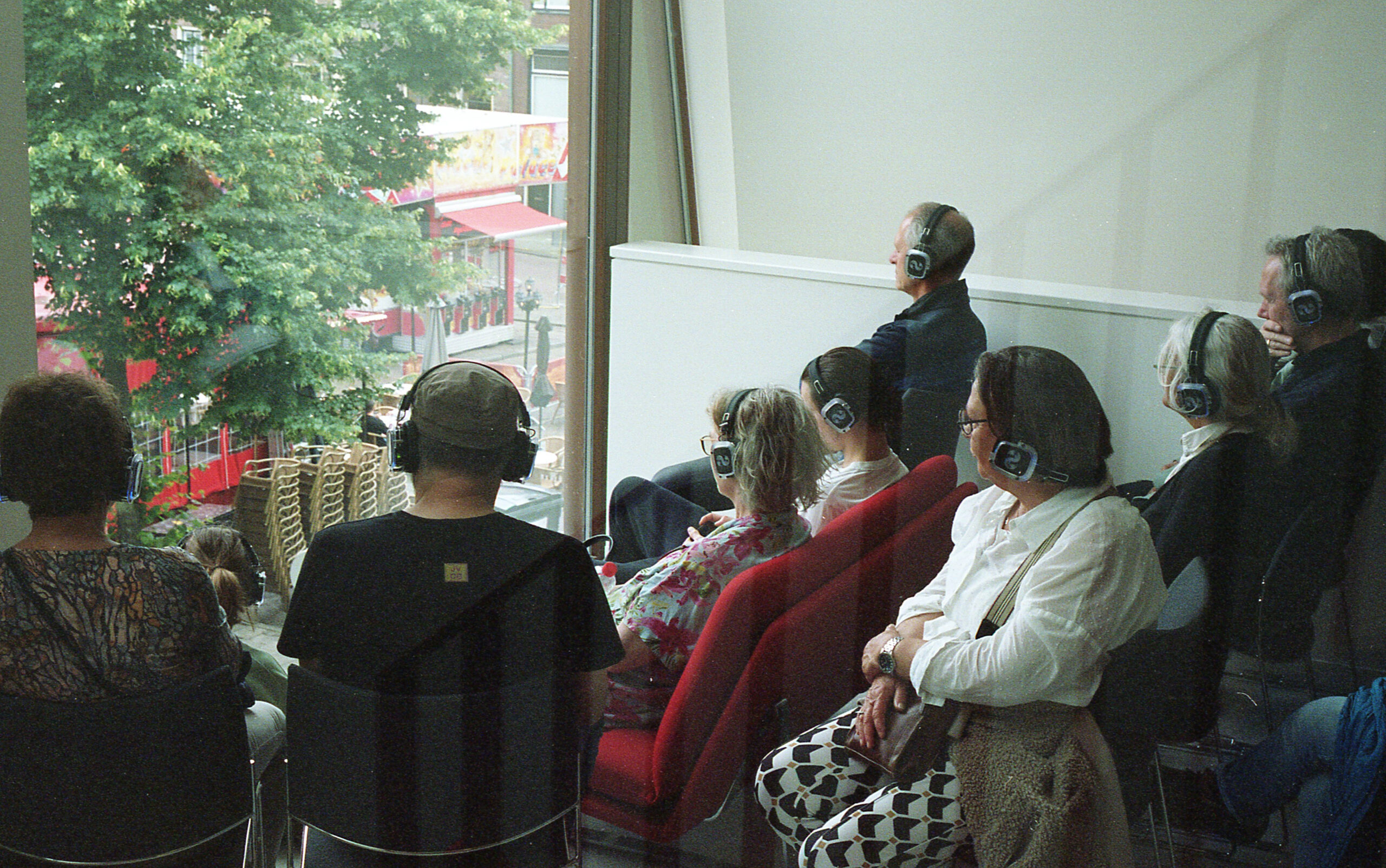 en groep mensen zit binnen in rijen stoelen en kijkt door een grote glazen ruit naar buiten. Alle aanwezigen dragen identieke grote, zwarte koptelefoons, wat suggereert dat ze naar dezelfde audio luisteren. Buiten het raam zijn groene bomen zichtbaar en een straat of plein met onder andere een rode luifel.